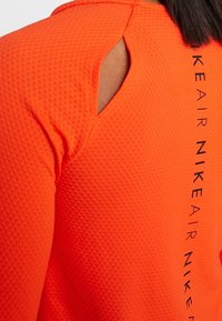 Orange atletisk skjorte med et struktureret mønster, åben skulderdesign og sort branding langs sidesømmen.