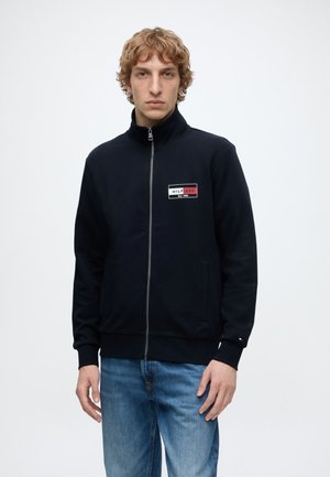 Junger Mann trägt eine schwarze Jacke mit Reißverschluss und einem "HILFIGER" Logo-Patch auf der Brust sowie blaue Jeans vor einem schlichten weißen Hintergrund.