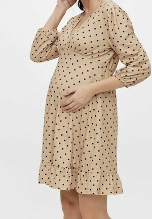 Kobieta w ciąży, ubrana w beżową sukienkę ciążową w czarne groszki, z rękawami 3/4 o fasonie bufek i falbaną u dołu, z ręką spoczywającą na brzuchu.
