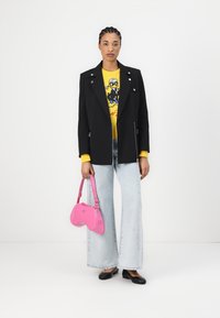 Blazer nero con hardware argento, maglione grafico giallo, jeans a gamba larga azzurri chiari e borsa rosa, completato con ballerine nere.