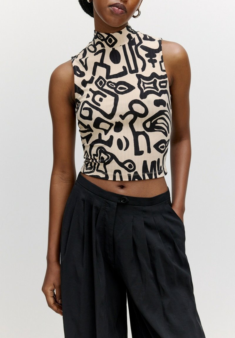 Femme portant un crop top beige sans manches avec des formes noires abstraites et un pantalon noir ample taille haute, debout devant un fond uni.