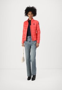 Padded coral jacket z zamkiem błyskawicznym i stojącym kołnierzem, zestawiona z jasnoniebieskimi dżinsami i czarnymi botkami na obcasie. Trzyma małą białą torbę.