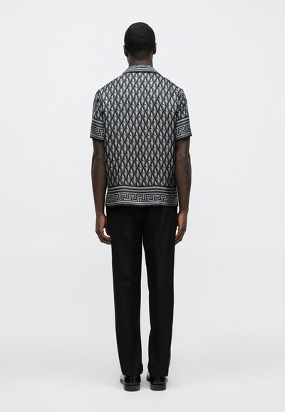 Μαύρο κοντομάνικο πουκάμισο με γεωμετρικά λευκά μοτίβα, αντίθετο οριζόντιο περιθώριο, φορεμένο με μαύρο παντελόνι, απεικονίζεται από πίσω.