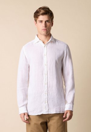 Jonge man met lichtbruin haar, gekleed in een wit knoopoverhemd met lange mouwen en beige broek, staande tegen een effen lichte achtergrond.