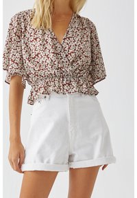 Femme portant un short blanc à ourlet roulé et un chemisier à manches courtes à motifs floraux avec taille volantée, debout devant un fond blanc uni.
