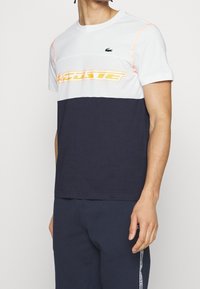 Kortärmad t-shirt med vit överdel, marinblå nederdel och meshpanel med orange logotyp. Lacoste-logotyp på bröstet, tillverkad av bomull.