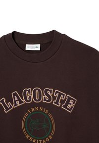 Sudadera de algodón marrón con lettering en blanco y amarillo "LACOSTE", logo de herencia de tenis en verde con un cocodrilo y cuello redondo.