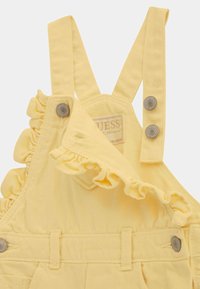 Pantalones de mezclilla amarillos con detalles de volantes, tirantes ajustables y botones a presión. Presenta una etiqueta de marca en la parte delantera.