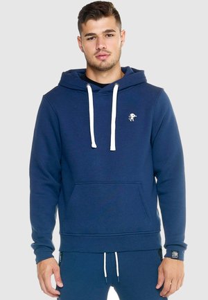 Giovane uomo che indossa una felpa con cappuccio blu navy con cordini bianchi e pantaloni della tuta blu navy abbinate su uno sfondo semplice.