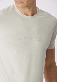 T-shirt in cotone beige chiaro con scollo rotondo. Logo "USPA SPORT" goffrato al centro, maniche corte e texture morbida.