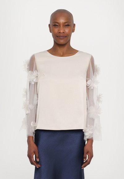 Woman wearing a cream top with sheer sleeves adorned with 3D floral appliques and a navy blue satin skirt, standing against a plain background.