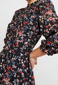 Robe fleurie en tissu noir avec des motifs rouges, bleus et blancs. Elle dispose d'un col volanté et de poignets froncés, ainsi qu'une taille ceinturée.