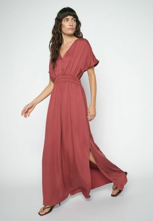 Kvinde iført en rust-rød maxikjole med taljeret pasform og korte ærmer, gående, med en sidelomme og iført sorte sandaletter med hæl.