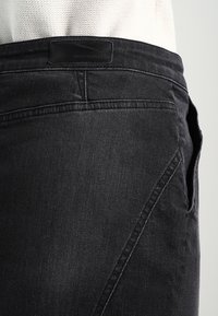 Jeans de mezclilla negra con una costura lateral curva, textura sutil y una etiqueta de marca de cuero en la cinturilla.