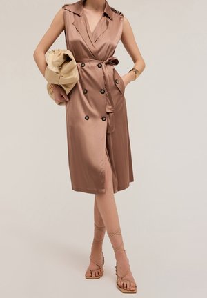 Satin, sleeveless trench dress in light brown with double-breasted buttons, belted waist, side pockets, and a thigh-high slit. Styled with sandals.