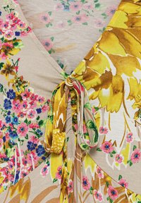 Floral print garment featuring a beige background with multicolored flowers in pink, yellow, and green; tied accent at the neckline.