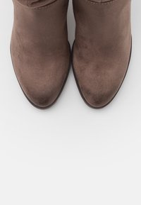 Bottes chevilles en daim marron avec un bout pointu et une texture lisse, présentant un détail discret de couture le long du bord supérieur.