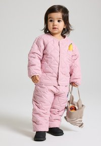 Vaaleanpunainen tikattu takki ja housut, joissa on teksturoitu pinta ja tasku, joka sisältää banaanin. Mustat saappaat ja beige laukku, jossa on hedelmiä.