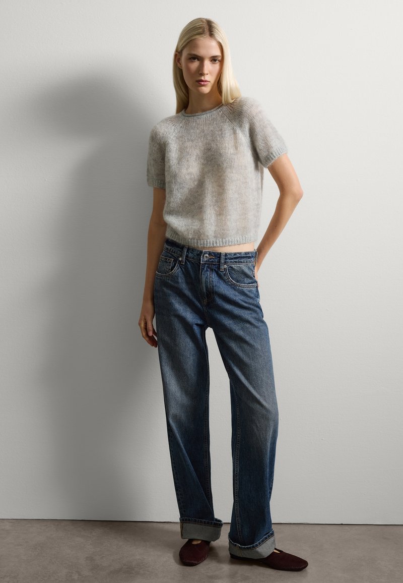 Donna bionda che indossa un maglione grigio corto e morbido a maniche corte, jeans blu larghi arrotolati alle caviglie e scarpe nere basse, in piedi contro un muro semplice.