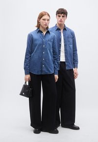 Jeans srajce v srednje modri barvi z gumbi spredaj, kombinirane s črnimi hlačami širokih nog. Modelka drži majhno črno torbico in stoji pred enobarvnim ozadjem.