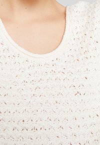 Biała dzianinowa bluzka z fakturowanym wzorem, okrągłym dekoltem oraz miękkim, lekkim materiałem z widocznymi detalami szwów.