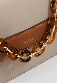 Bolso cruzado de imitación de cuero en color beige y marrón, con una cadena de tortuga gruesa y accesorios en dorado, además de detalle del logo en relieve.