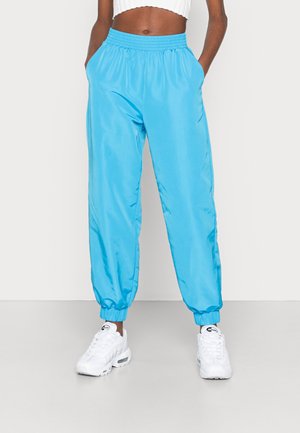 Personne portant un pantalon bleu vif avec taille élastique et revers, des baskets blanches et un haut court blanc côtelé, debout les mains dans les poches.