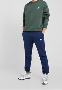 Nike grön sweatshirt med vitt logo, i kombination med marinblå joggers med vitt logo. Modellen bär grå sneakers med mönstrade detaljer.