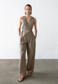 Beżowa lniana bluzka typu wrap z głębokim dekoltem w kształcie V, zestawiona z szerokimi spodniami. Lekka tekstura, luźny krój, z dodatkowym akcentem w postaci wiązania. Czerwone klapki.