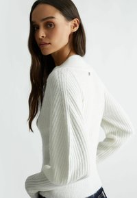 Maglione bianco a costine con colletto alto e texture diagonale prominente. Presenta un piccolo dettaglio con logo sul retro.