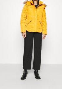 Giacca imbottita gialla con cappuccio e bordo in pelliccia sintetica, tasche con zip e texture trapuntata; abbinata a pantaloni larghi neri e stivaletti.