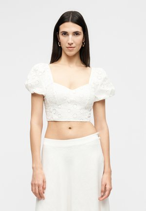 Moteris su ilgais tamsiais plaukais, vilkinti baltą trumpą palaidinę su akių raštais ir pūstomis rankovėmis bei baltą sijoną, stovinti prieš paprastą foną.