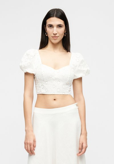 Femme aux longs cheveux foncés portant un chemisier court blanc en œillet avec des manches bouffantes et une jupe blanche, debout devant un fond uni.