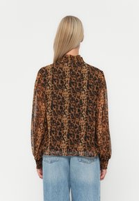 Prozirna smeđa bluzica s crnim uzorkom leopard, visoki nabran ovratnik i dugi rukavi. Uparena s svijetloplavim trapericama visokog struka.