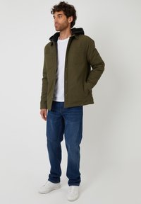 Olivgrön jacka med svart huva, två bröstfickor, knappstängning, bärs över en vit t-shirt och parat med blå jeans och vita sneakers.