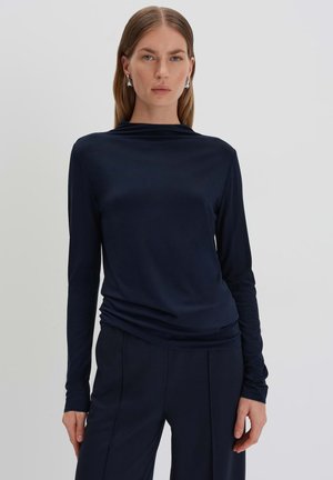 Langärmliges, dunkelblaues Oberteil mit drapiertem Ausschnitt und glattem Stoff. Kombiniert mit maßgeschneiderter dunkelgrauer Hose, die ein minimalistisches Design aufweist.