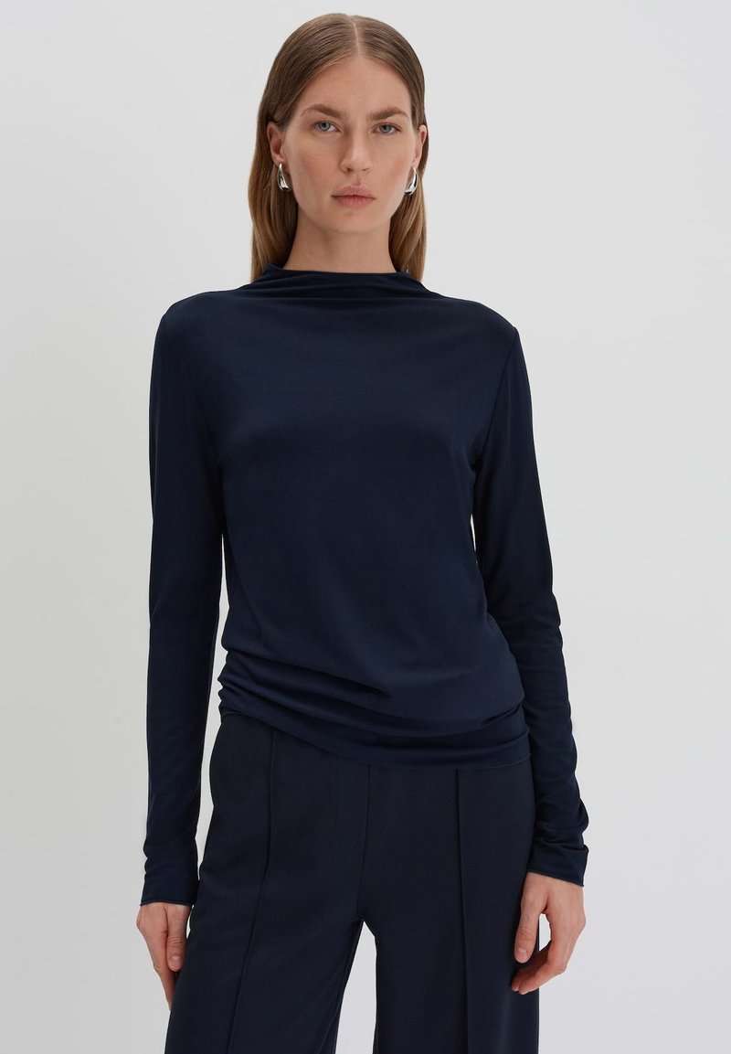Langärmliges, dunkelblaues Oberteil mit drapiertem Ausschnitt und glattem Stoff. Kombiniert mit maßgeschneiderter dunkelgrauer Hose, die ein minimalistisches Design aufweist.