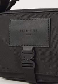 Black textured bag featuring a prominent logo patch, a sturdy clasp closure, and flat straps. Material appears durable and water-resistant.