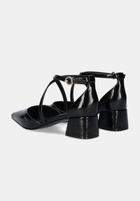 Zapatos de charol negro con tacón cuadrado, punta afilada y correas entrelazadas. Presentan un acento de hebilla dorada en la correa del tobillo.