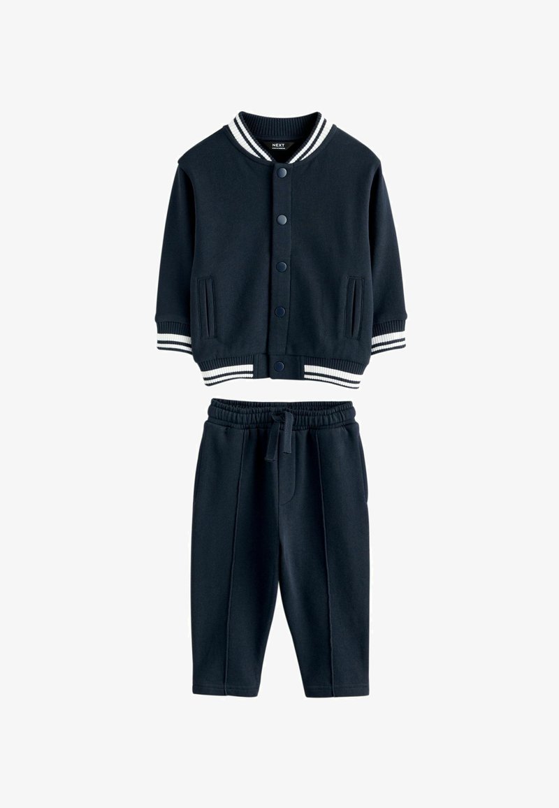 Tamsiai mėlynas džemperis su gumuotu apykakle ir dryžuotomis rankovėmis, derinamas su atitinkamais elastiniais kelnais su priekiniu siūlu.