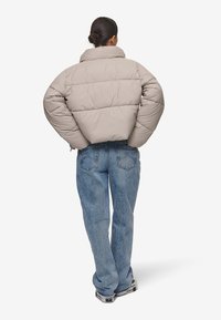 Beige pufferjakke med høy krage og boksform. Den har horisontale sømmer og er brukt over løse blå denimjeans.