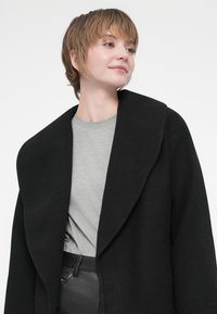 Casaco preto oversized com lapelas largas, usado sobre uma camisa cinza-clara. O casaco tem uma textura suave e está desabotoado, revelando a camisa por baixo.