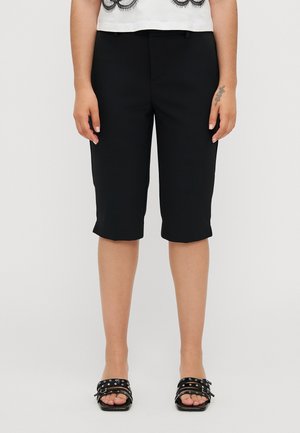 Svarte kne-lange shorts med en glatt stoff, med en skreddersydd design og sidelommer. Kombinert med svarte sandaler med stropp.