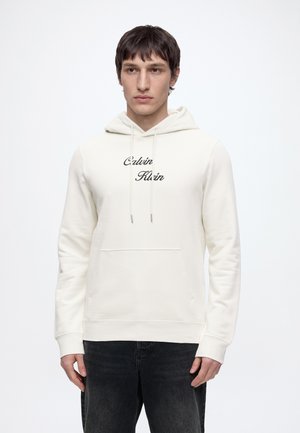Młody mężczyzna ubrany w białą bluzę z kapturem Calvin Klein z przednią kieszenią i czarne dżinsy, stojący na jednolitym jasnym tle.