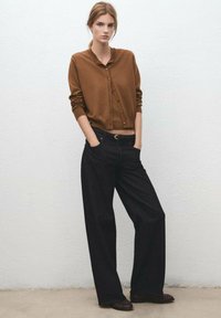 Cárdigan marrón de botones, hecho de una tela suave, combinado con jeans de pierna ancha de talle alto en negro. La modelo está de pie contra un fondo de pared neutro.