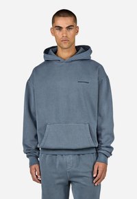 Hettegenser i falmet blå bomull med lommes foran, ribbestrikkede mansjetter og brodert logo på brystet. Jevn tekstur.