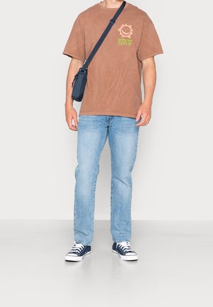 Mand iført brun grafisk t-shirt, lyseblå jeans, marineblå sneakers og en sort tværskuldertaske, der står op ad en ensfarvet væg.