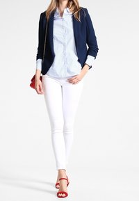 Mujer caminando con un blazer navy, una camisa de rayas azules, pantalones skinny blancos, tacones rojos y llevando un pequeño bolso rojo sobre su hombro.