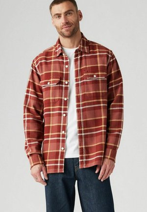 Camisa de franela en rojo con un patrón a cuadros amarillo y blanco, que presenta dos bolsillos en el pecho y botones blancos, llevada sobre una camiseta blanca.