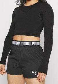 Musta pitkähihainen crop top, jossa on teksturoitu muotoilu ja Puma-logo; yhdistetty mustiin shortseihin, joissa on valkoinen Puma-brändätty vyötärö.